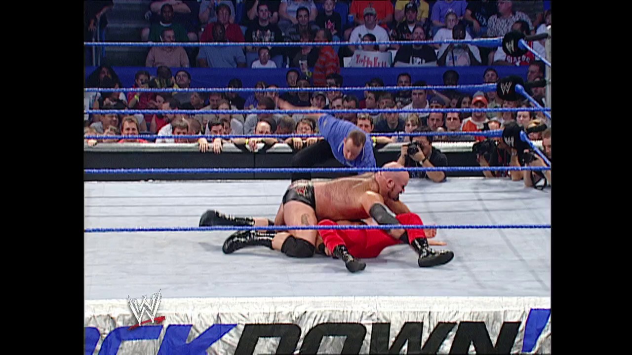 A frame from WWE SmackDown (September 4, 2003) at 23:58. A wrestler in red attire lies on the mat while another in dark trunks applies a submission hold during a WWE SmackDown match. The blue-roped ring is surrounded by a packed crowd, with the WWE logo visible at the bottom of the frame.