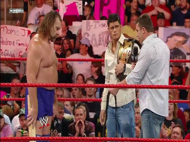 A frame from WWE Raw (July 14, 2008) at 39:00. A WWE wrestler stands shirtless in the ring wearing blue pants while two men in casual attire hold a championship belt, with an enthusiastic crowd visible in the background behind the red ropes.