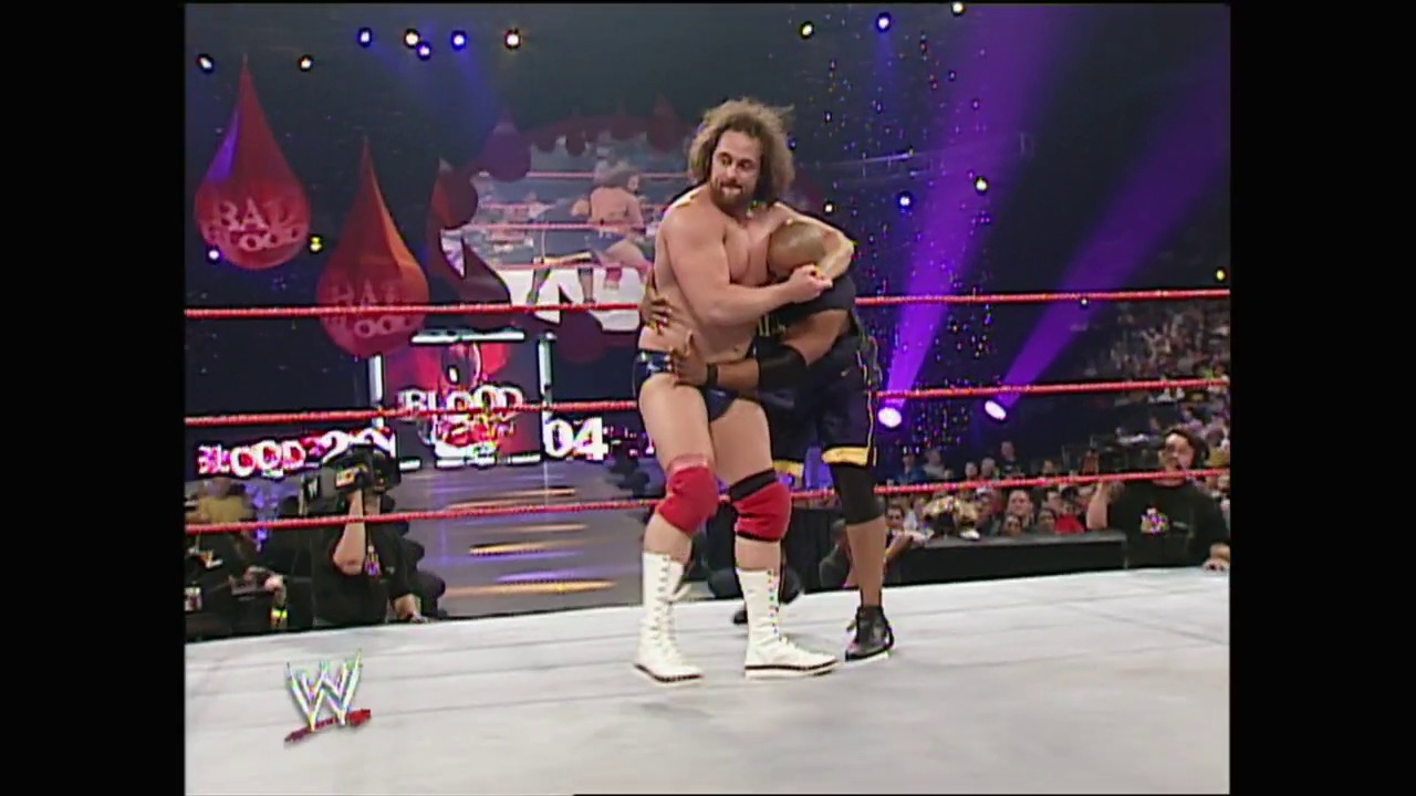 A frame from WWE Bad Blood (2004) at 1:12:33. A WWE wrestler with long blonde hair performs in the ring during Bad Blood 2004, executing a move on his opponent. The red-roped wrestling ring is surrounded by a crowd, with purple and red stage lighting and WWE graphics visible in the background.