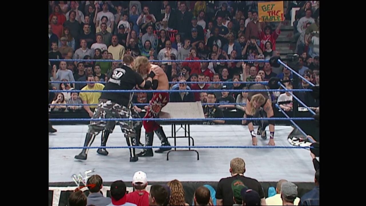 A frame from WWE SmackDown (March 22, 2001) at 1:02:55. Two wrestlers engage in combat inside a blue-roped WWE ring with a folding chair nearby, while a packed crowd watches from the stands. A "GET THE TABLE" sign is visible in the background among the enthusiastic audience members.