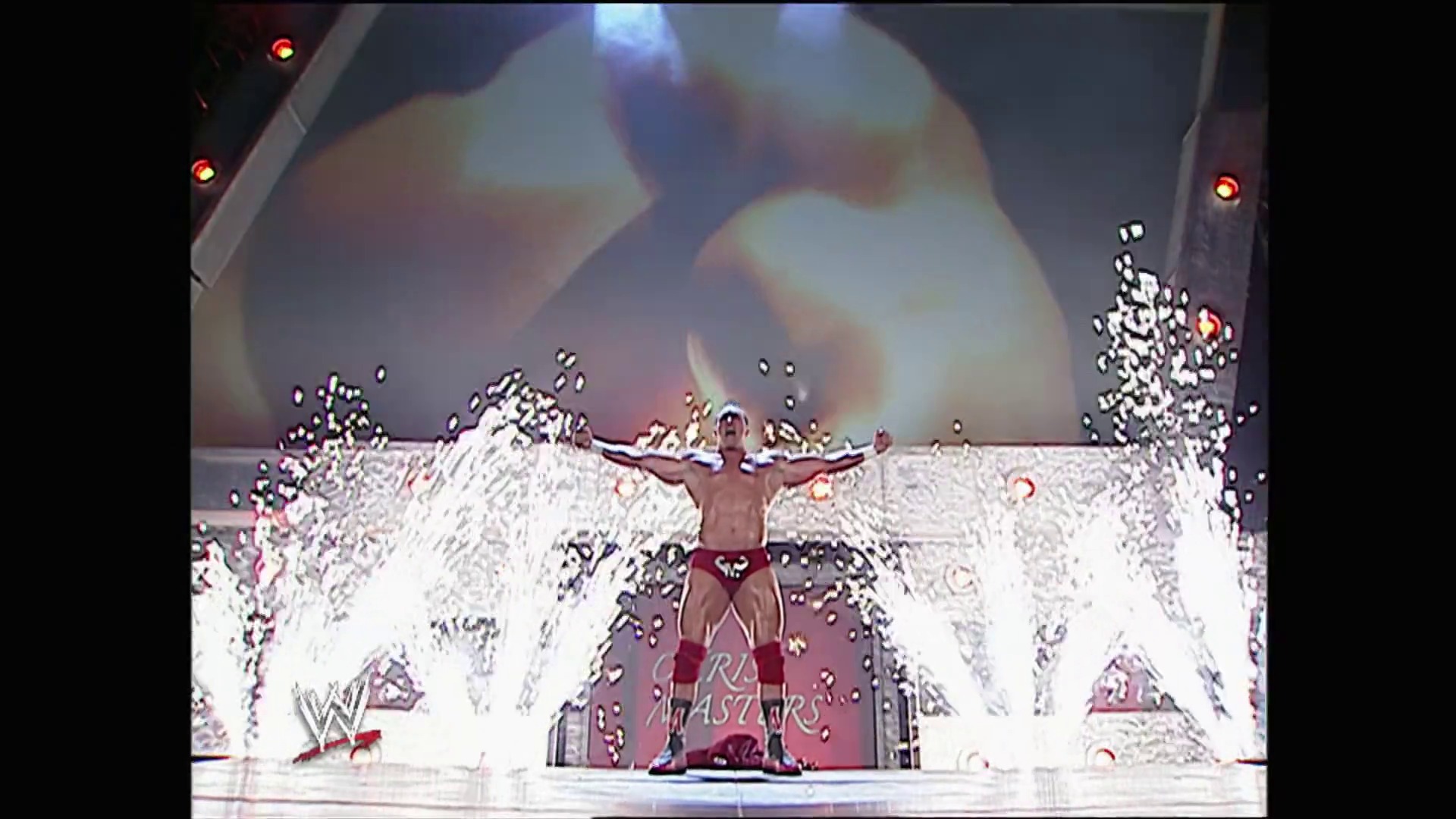 A frame from WWE Raw (January 9, 2006) at 38:40. A shirtless wrestler stands triumphantly with arms raised on an elaborate entrance stage featuring red and white pyrotechnics exploding around him. The WWE logo is visible on the stage set, with dramatic lighting and visual effects creating an intense atmosphere typical of professional wrestling broadcasts.
