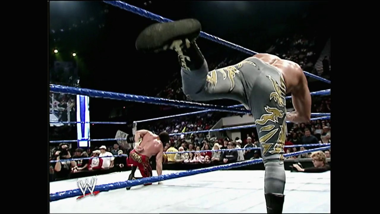 A frame from WWE No Way Out (2004) at 1:03:59. A WWE wrestler in gray and gold patterned pants performs an aerial move over a downed opponent in red in the ring, with the blue ropes visible and an engaged crowd in the background during the No Way Out event.