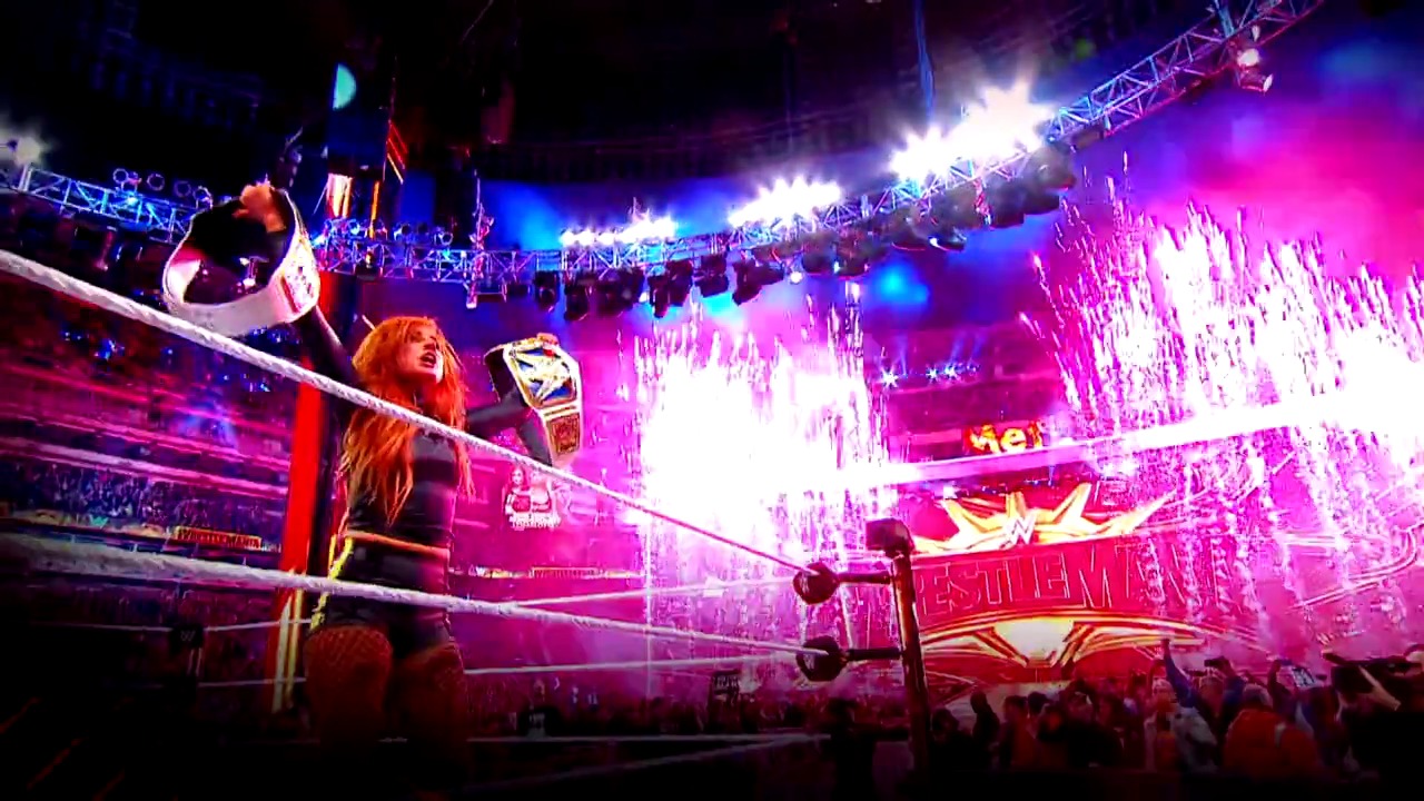 A frame from WWE Royal Rumble (2022) at 1:01. A wrestler with red hair stands in a WWE ring holding a championship belt, surrounded by vibrant pink and blue lighting effects. The arena is filled with spectators and large illuminated graphics displaying "WrestleMania" branding overhead.