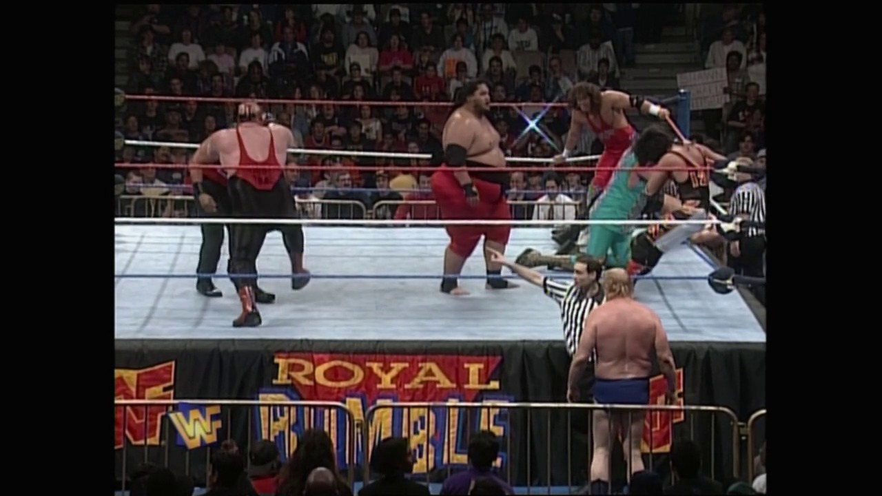 A frame from Royal Rumble (1996) at 1:22:17. A multi-wrestler match unfolds in a WWF ring during Royal Rumble 1996, with competitors in red and teal attire grappling while referees oversee the action. The crowd fills the arena seats in the background as event signage is visible along the ring apron.