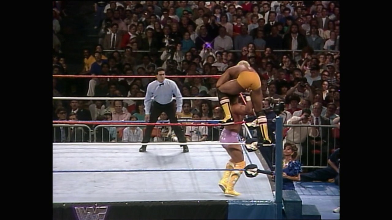 A frame from WrestleMania IV (1988) at 1:01:01. A wrestler in yellow and black attire performs an aerial maneuver in a WWF ring during WrestleMania IV, with a full crowd visible in the background and an official in a light blue shirt overseeing the match.