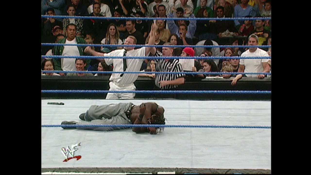 A frame from WWE SmackDown (November 23, 2000) at 37:10. A wrestler lies prone on the mat in a WWE SmackDown ring, with a referee in striped shirt standing nearby and a packed crowd visible in the background. The blue ropes frame the scene, and the WWF logo is visible in the lower left corner.