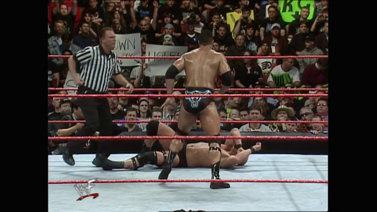 A frame from WrestleMania XV (1999) at 2:22:28. A referee in striped shirt stands in a WWE wrestling ring as one shirtless wrestler lies on the mat while another stands nearby, with an enthusiastic crowd visible in the background behind the red ropes.