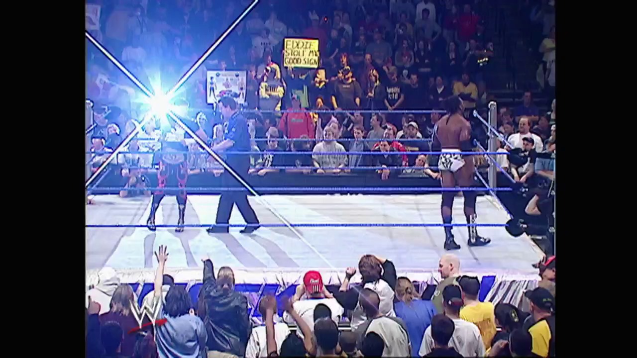 A frame from WWE SmackDown (March 25, 2004) at 1:13:56. A professional wrestling match unfolds in a packed arena, with wrestlers in the ring and an engaged crowd in the foreground. Bright stage lighting illuminates the blue-roped ring while spectators fill the stands and ringside area.