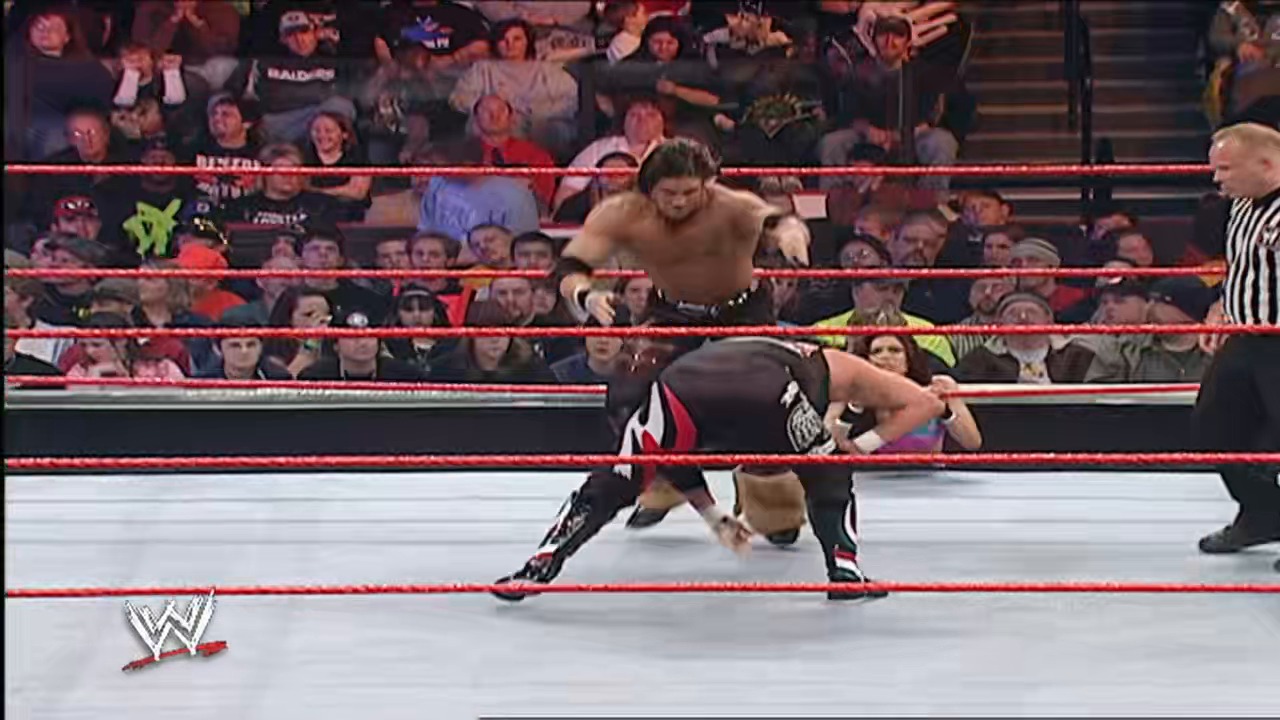 A frame from WWE Raw (February 5, 2007) at 38:28. A WWE wrestler in tan trunks stands in the red-roped ring while another competitor in black and red attire crouches low during an offensive move, with a referee in striped shirt nearby and a crowd visible in the background.