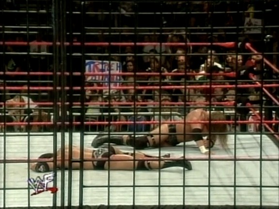 A frame from WWF Rebellion (1999) at 2:03:12. A wrestler lies prone on the mat inside a steel cage during a WWF match, with the crowd visible in the bleachers behind the cage structure and the WWF logo displayed on the ring apron.
