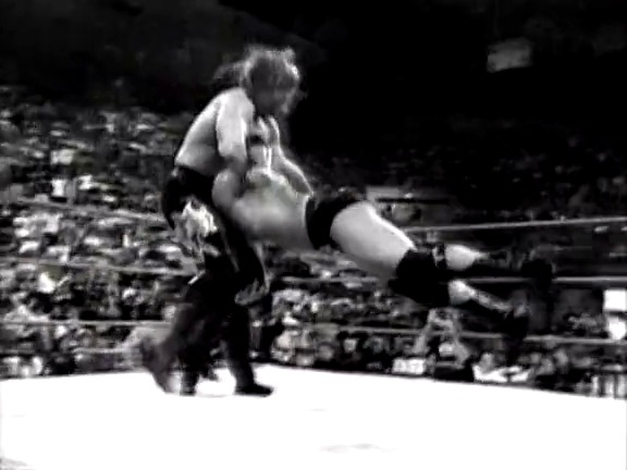 A frame from WWF Rebellion (1999) at 0:45. A wrestler executes a move on their opponent in the ring during a WWF match, with a crowd visible in the blurred background. The black and white footage captures the dynamic action typical of professional wrestling events from this era.