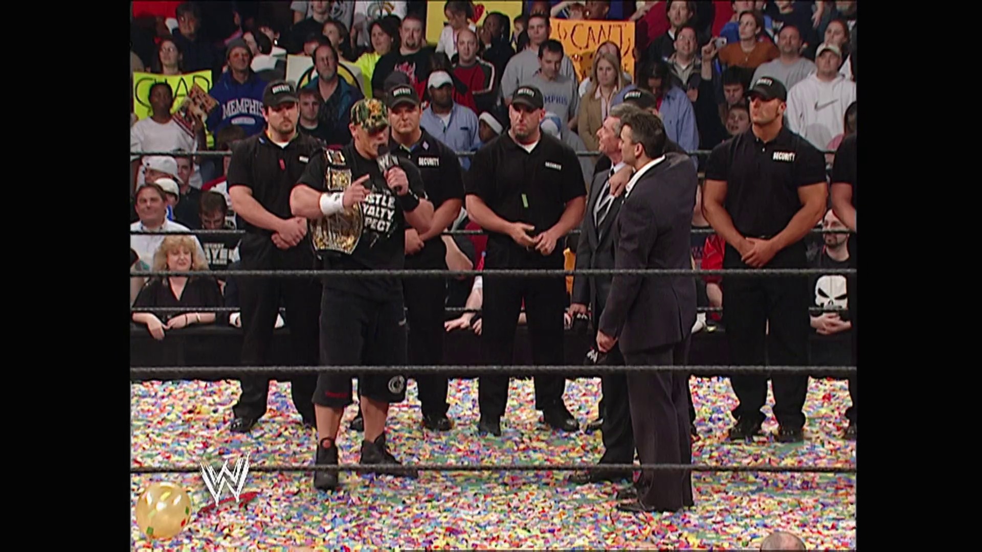 A frame from WWE Raw (March 20, 2006) at 13:15. A WWE wrestling ring is surrounded by colorful confetti during a celebratory moment, with multiple wrestlers in black shirts standing in the ring alongside a suited official. The crowd fills the arena in the background, with various signs visible in the stands.