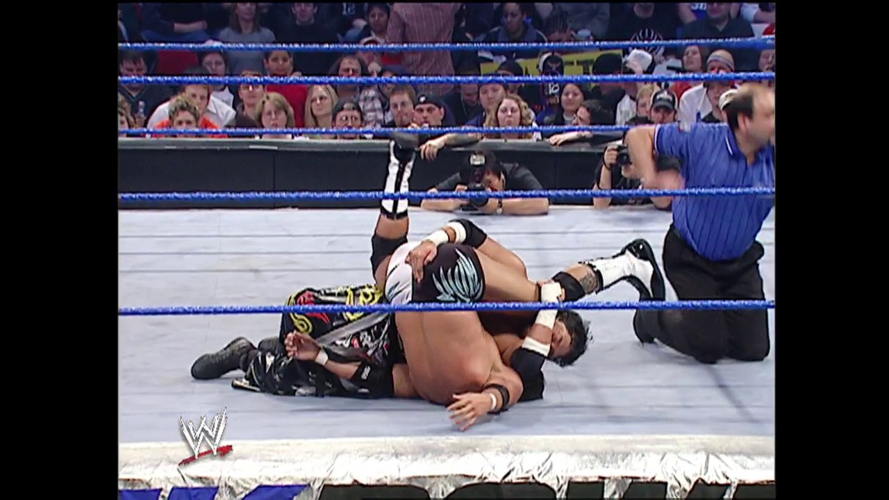 A frame from WWE SmackDown (January 22, 2004) at 22:25. # WWE SmackDown Match Scene

Two wrestlers are engaged in a match on the blue-roped WWE ring, with one wrestler pinned down while another stands nearby. The crowd fills the arena seats in the background, and a referee in a blue shirt oversees the action. The WWE logo is visible in the bottom left corner.