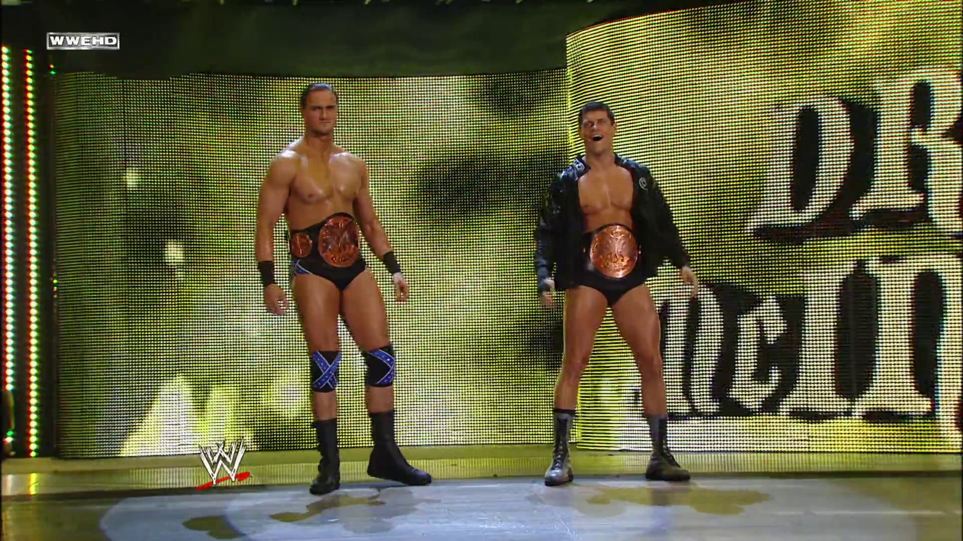 A frame from WWE Raw (September 20, 2010) at 13:20. Two wrestlers wearing championship belts stand on the WWE Raw ring against a bright yellow LED screen backdrop. The wrestler on the right wears a black jacket over his singlet, while both display their bronze championship belts.