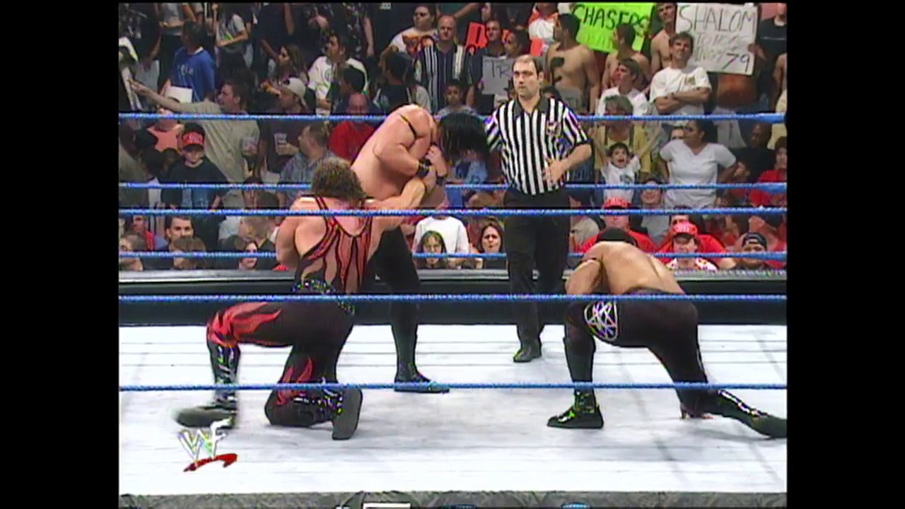 A frame from WWE SmackDown (September 14, 2000) at 26:47. A WWE SmackDown wrestling match in progress features two wrestlers in the ring—one shirtless in red and black attire positioned above another wrestler in black pants. A referee in striped shirt stands nearby as a crowd watches from the stands behind the blue-roped ring.