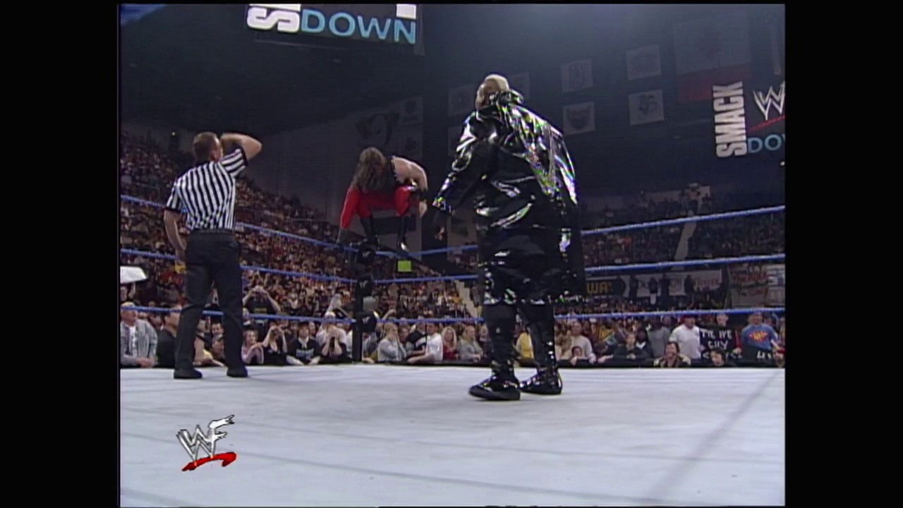 A frame from WWE SmackDown (November 18, 1999) at 38:24. A WWE SmackDown match in progress features a wrestler in black and neon attire standing in the ring, while a referee in striped shirt and another competitor in red observe. The packed arena crowd is visible in the background with "DOWN" signage visible at the top.