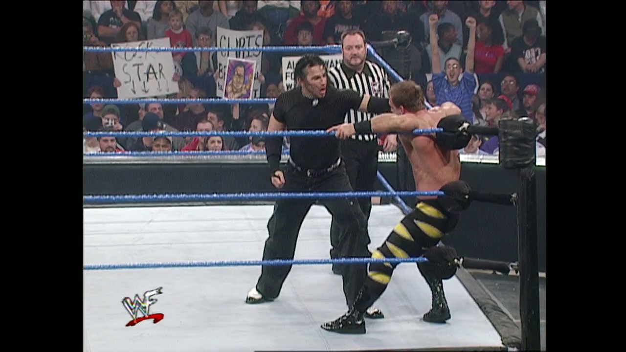 A frame from WWE SmackDown (December 28, 2000) at 13:39. A WWE wrestler in black and yellow attire grapples with an opponent in the ring while a referee in striped shirt supervises. The crowd fills the arena stands behind the blue-roped ring, with visible fan signs and the WWF logo displayed at ringside.