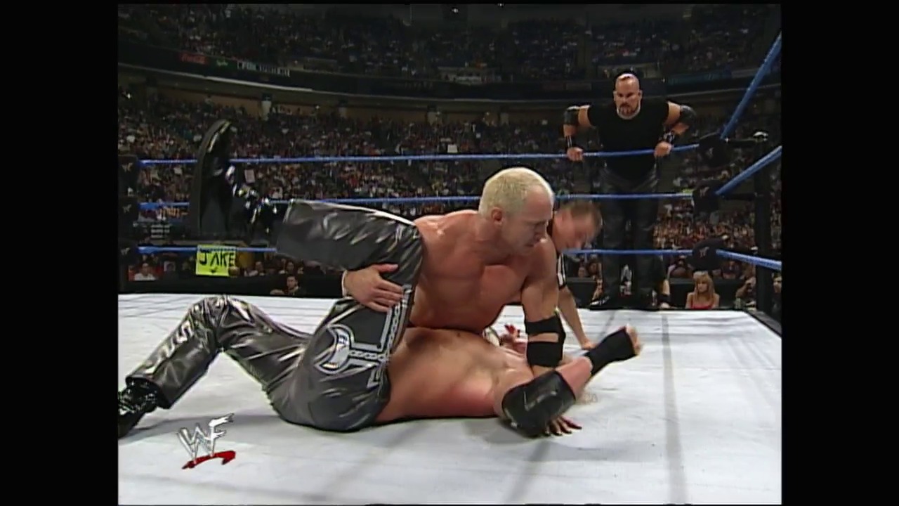 A frame from WWE SmackDown (April 27, 2000) at 38:14. A WWE wrestler in black and silver pants lies on the mat during a match, while another competitor applies pressure. The referee stands nearby as the crowd fills the arena in the background, with the distinctive blue ring ropes and WAF logo visible.