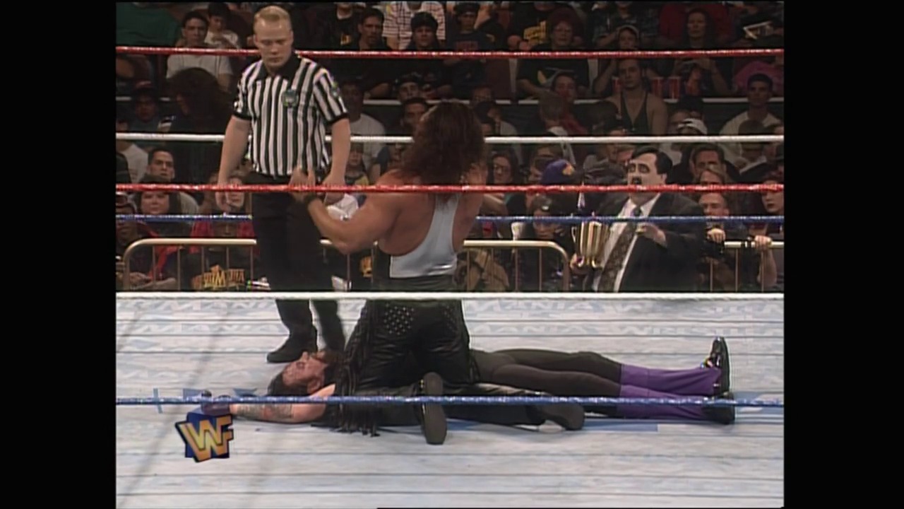 A frame from WrestleMania XII (1996) at 1:11:58. A WWF wrestler lies on the mat after being struck, while an opponent stands nearby and a referee in a striped shirt oversees the match. The crowd fills the arena seats in the background, with the WWF logo visible on the ring apron.