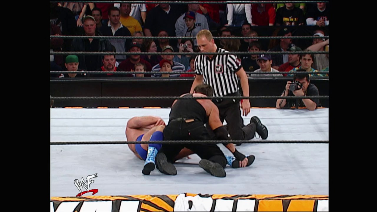 A frame from Royal Rumble (2002) at 46:45. A wrestler lies on the white mat while a referee in black and white stripes stands over him during a WWE Royal Rumble match, with the crowd visible in the background behind the ropes.