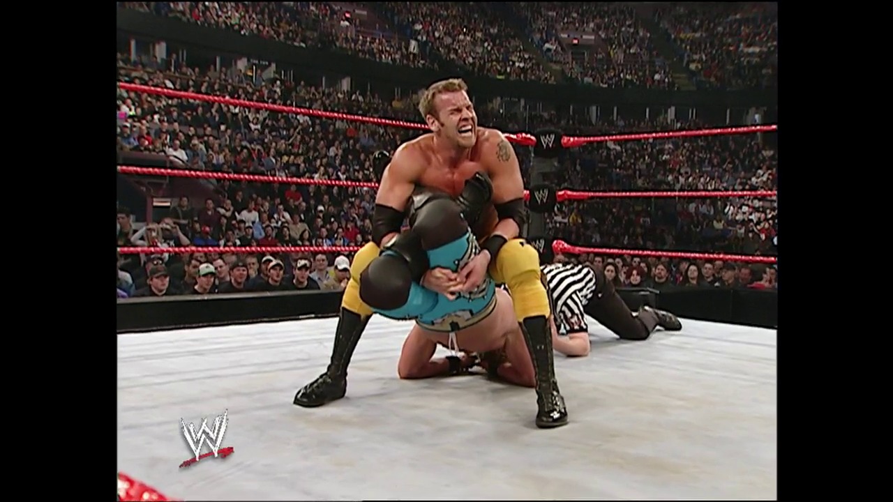 A frame from WWE Backlash (2004) at 45:01. A WWE wrestler in black and yellow arm sleeves performs a move in the ring during a televised match, with the packed arena crowd visible in the background and the distinctive red WWE ropes framing the action.