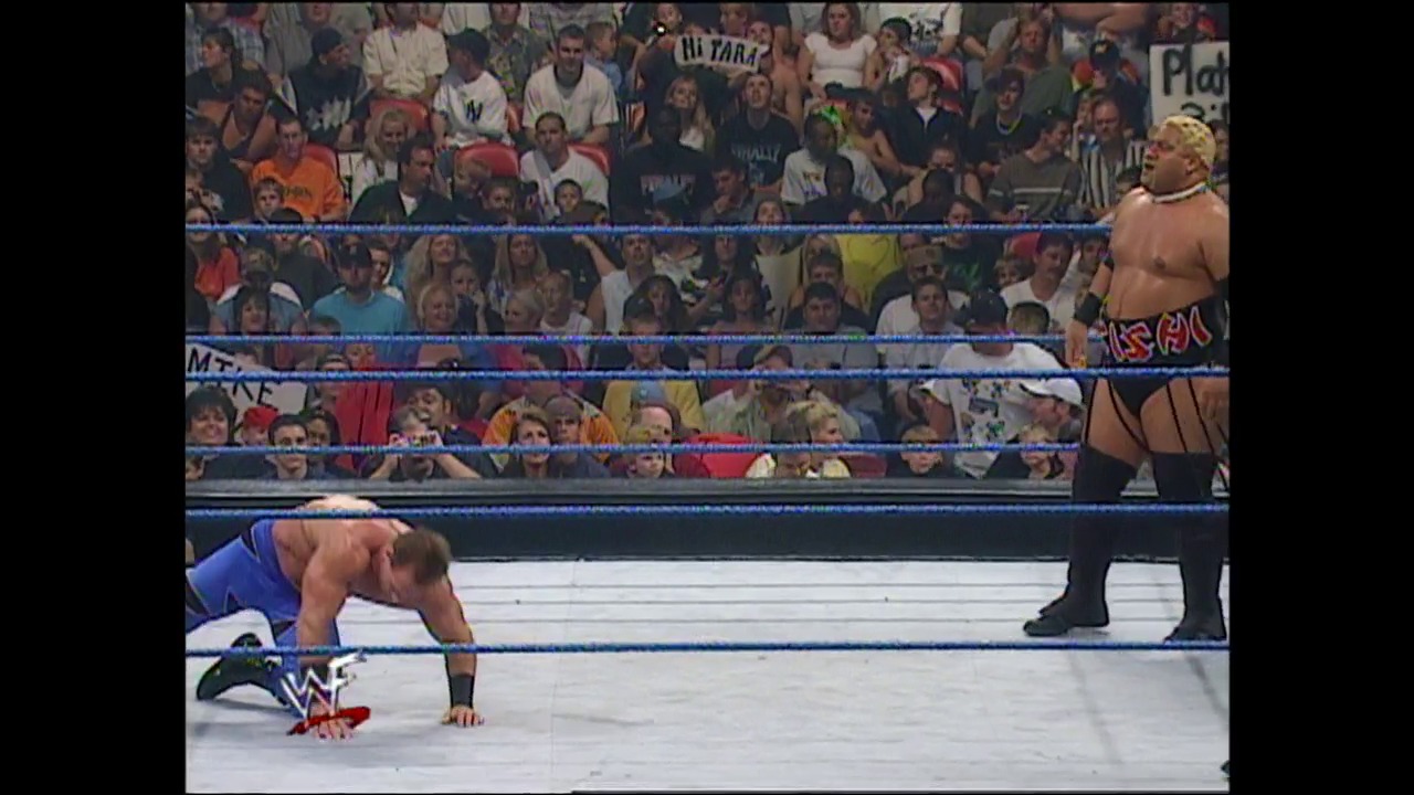 A frame from WWE SmackDown (September 7, 2000) at 26:00. A WWE wrestler in blue trunks is on the mat in a prone position inside the blue-roped ring, while another shirtless wrestler stands nearby. The crowd fills the arena stands in the background, with the WWE logo visible on the ring apron.