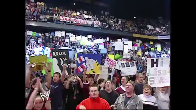 A frame from WWE SmackDown (January 6, 2005) at 1:00. A packed WWE SmackDown crowd fills the arena with colorful signs and enthusiasm, displaying various handmade placards and banners while seated in the bleachers during the live wrestling event.