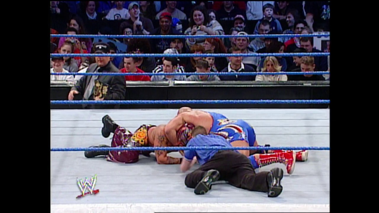 A frame from WWE SmackDown (January 23, 2003) at 1:01:59. A wrestler in blue attire lies on the mat during a WWE SmackDown match, with another performer nearby and the referee in position. The crowd watches from the stands behind the blue-roped ring, with the WWE logo visible in the lower left corner.