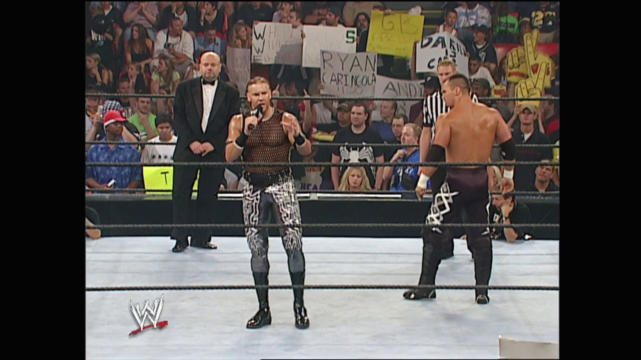 A frame from WWE Vengeance (2002) at 1:51:45. A WWE wrestler in distinctive black and white patterned pants stands in the ring holding a microphone, with a referee and another competitor visible. The crowd fills the stands behind the ropes with various signs, while the WWE logo appears in the lower left corner.