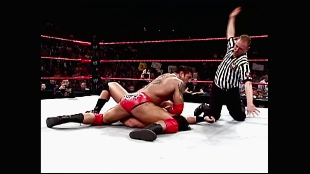 A frame from WWE Raw (January 3, 2005) at 0:49. A wrestler in red attire lies on the white mat of a WWE Raw ring while a referee in a striped shirt stands nearby with his arm raised. The ring is illuminated by red LED ropes, with a crowd visible in the darkened arena background.