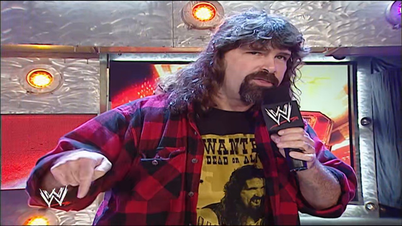 A frame from WWE Raw (June 11, 2007) at 37:11. A wrestler with long dark hair and a beard holds a microphone while wearing a red plaid shirt over a yellow graphic tee in a WWE ring set. Heat lamps glow above the metallic ceiling as the WWE logo appears in the corner of the frame.