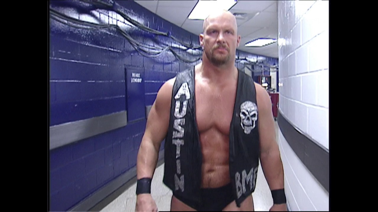 A frame from WWE SmackDown (November 9, 2000) at 0:51. A muscular bald wrestler stands in a backstage hallway wearing black wristbands and a skull-emblazoned black vest, looking directly at the camera with a serious expression. The blue-walled corridor behind him is typical of WWE's backstage area setup from that era.
