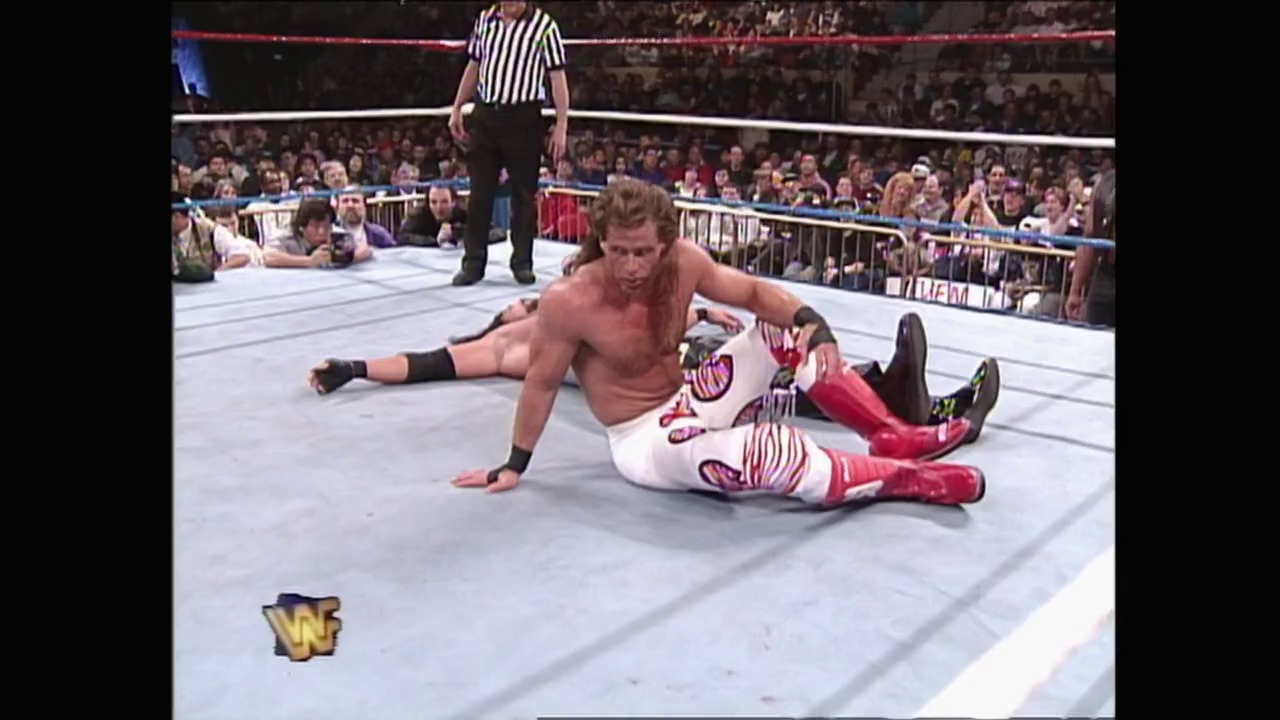A frame from WrestleMania XI (1995) at 1:48:29. A shirtless wrestler in red and white attire performs a submission move on an opponent in the WWF ring, while a referee in striped shirt oversees the match. The packed crowd lines the barriers as the WWF logo appears in the lower left corner.
