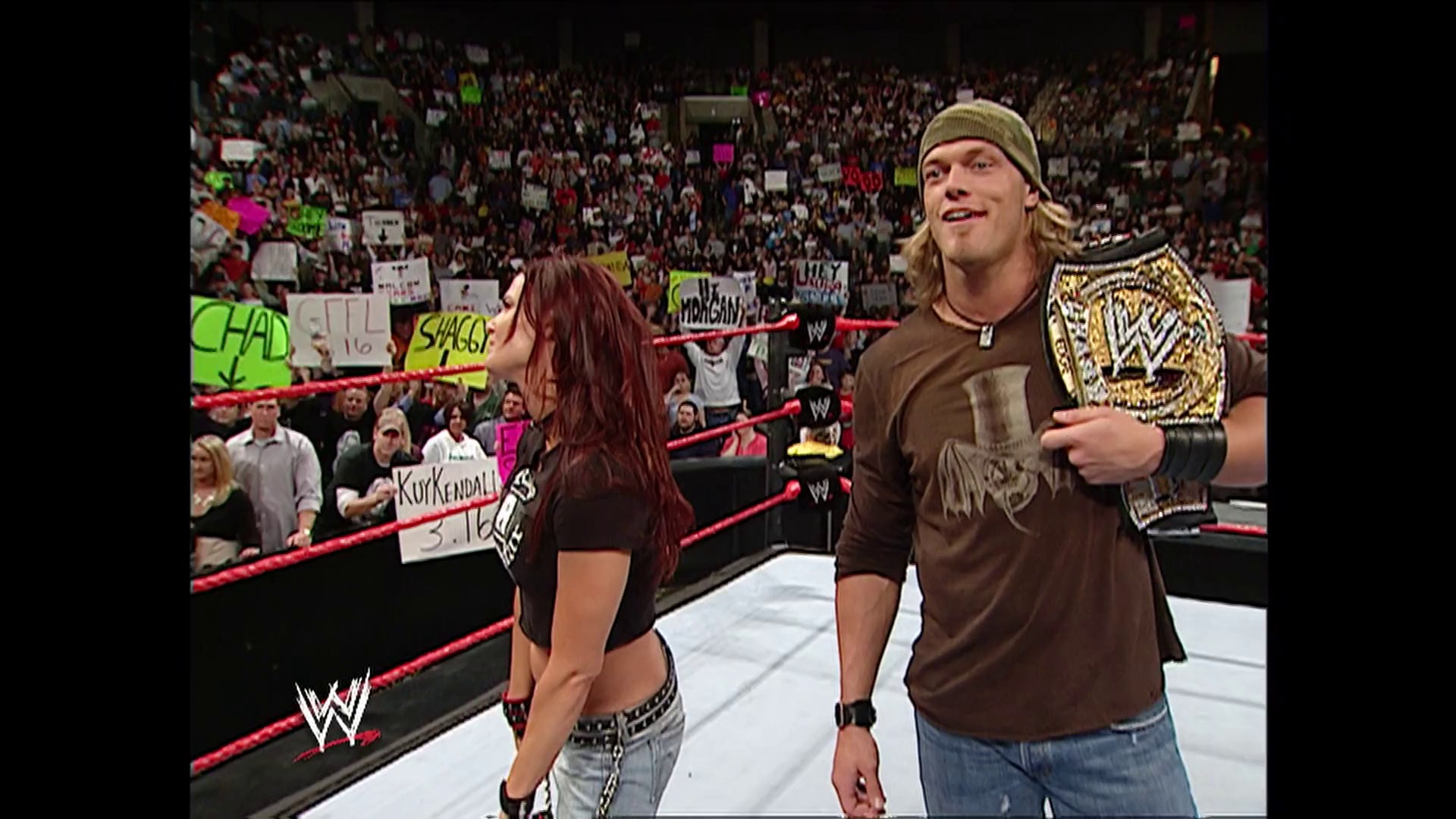 A frame from WWE Raw (January 23, 2006) at 14:25. # WWE Raw Ring Scene

A male wrestler in a brown shirt and beanie holds a championship belt while standing in the ring next to a female wrestler with reddish-brown hair. The crowd fills the arena behind the red ropes, holding colorful signs.