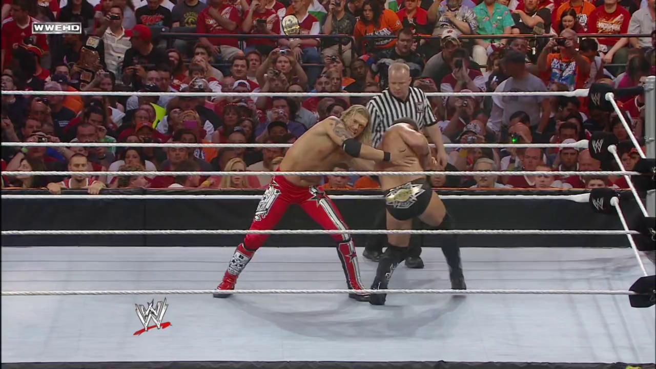 A frame from WrestleMania XXVII (2011) at 2:12:15. A WWE wrestler in red pants executes a move on an opponent in the ring, while a referee in black and white stripes officiates. The crowd fills the arena behind the ropes, with the WWE logo visible on the mat.