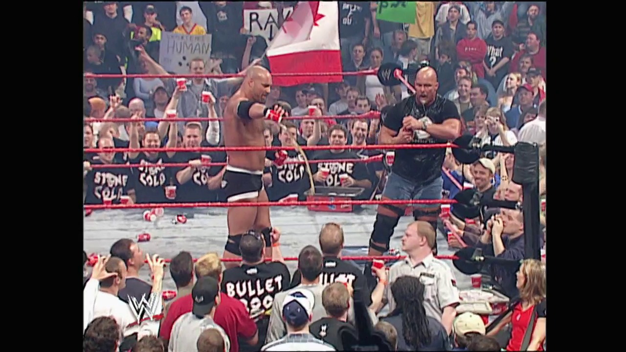 A frame from WWE Raw (May 5, 2003) at 1:34:56. # WWE Raw Match Scene

A shirtless wrestler stands in the red-roped ring while an official in black attends to ringside personnel. The crowd fills the arena around them, with Canadian flags visible in the background and WWE branding evident throughout the venue.