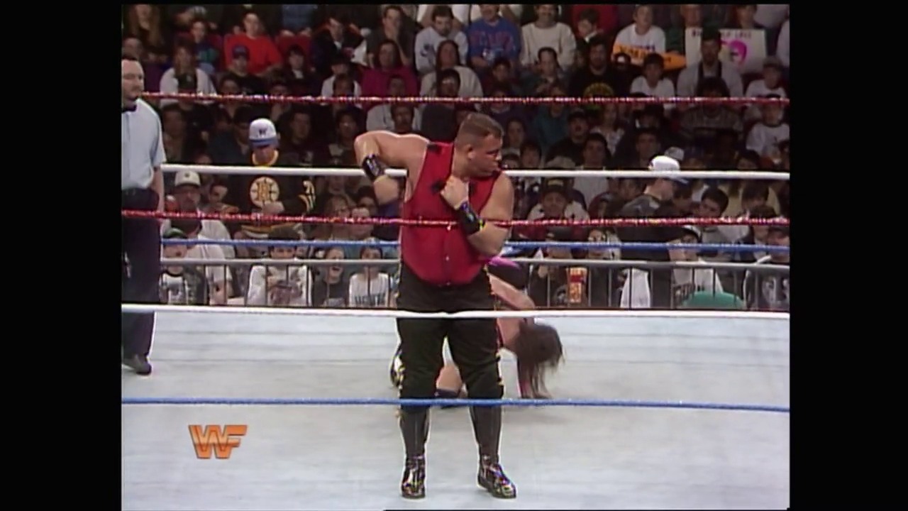 A frame from Royal Rumble (1994) at 23:25. A wrestler in a red tank top and black pants stands in the WWF ring before a packed crowd, with the iconic WWF logo visible on the mat canvas and the steel cage structure visible above.