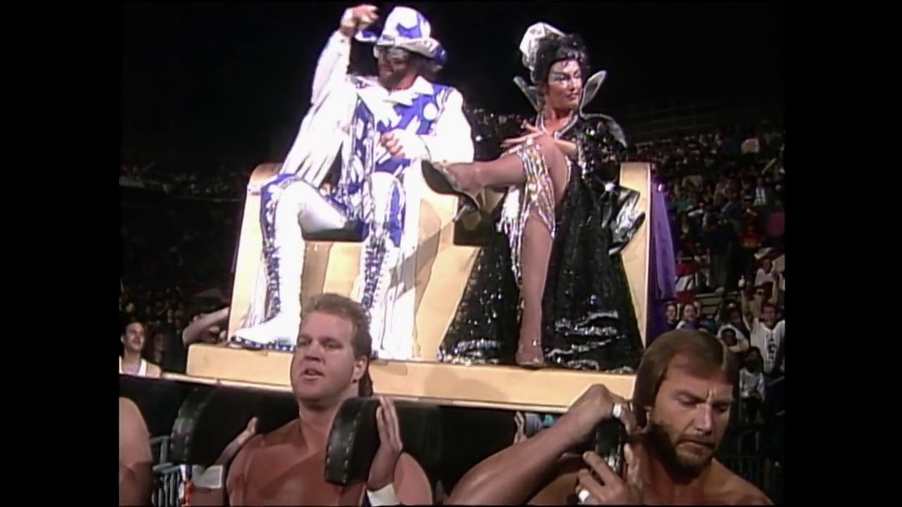 A frame from WrestleMania VII (1991) at 1:20:15. # WrestleMania VII Frame

Two wrestlers stand in the ring while an elaborately costumed performer sits elevated on the ropes, wearing a white and blue outfit with a decorative cape. The crowd is visible in the background of the wrestling arena.