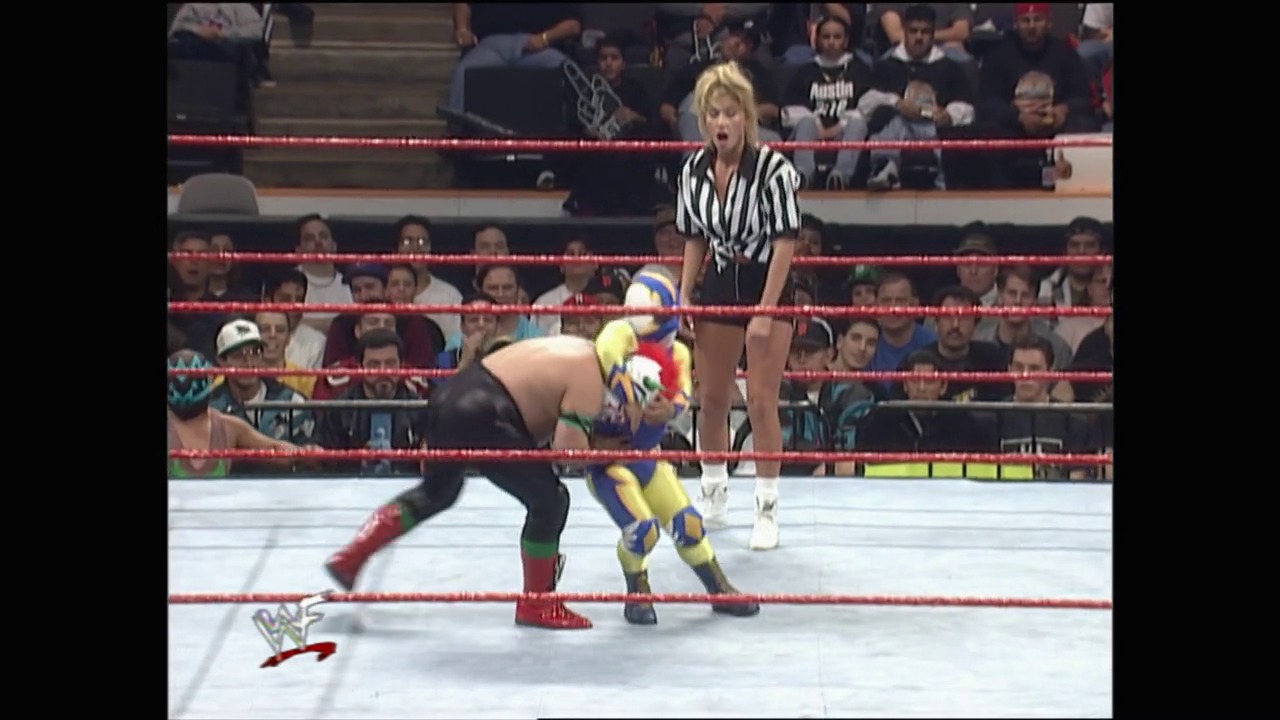 A frame from Royal Rumble (1998) at 23:29. A WWE wrestler in colorful blue and yellow attire grapples with an opponent in the ring while a referee in striped shirt oversees the match. The crowd fills the arena stands surrounding the red-roped wrestling ring.