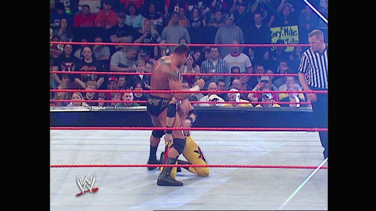 A frame from WWE Raw (February 14, 2005) at 50:10. A WWE wrestler in yellow and blue attire is on his knees in the ring during a match, with another wrestler standing nearby. The crowd fills the arena seats behind the red ropes, while a referee in a striped shirt observes the action.
