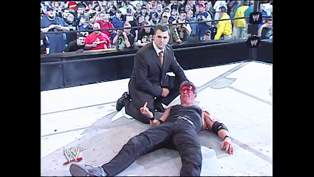A frame from WrestleMania XIX (2003) at 2:41:29. A WWE wrestler lies bleeding on the mat while a suited official kneels beside him during WrestleMania XIX. The packed crowd watches intently from the stands surrounding the white wrestling ring.