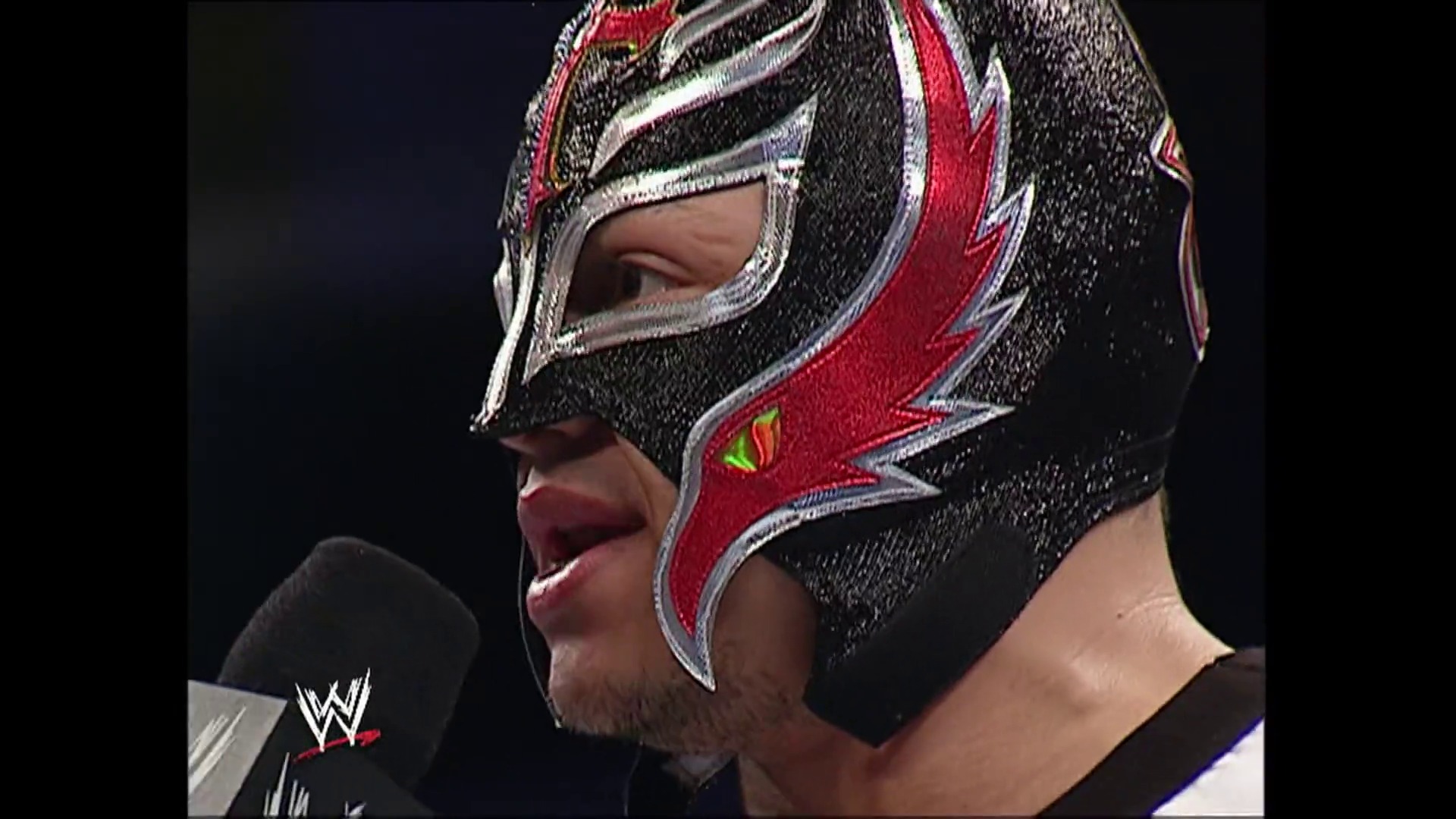 A frame from WWE SmackDown (March 31, 2006) at 11:56. A masked wrestler wearing a black and red sparkly lucha libre-style mask with silver trim speaks into a microphone during a WWE SmackDown event, captured against a dark background with the WWE logo visible.
