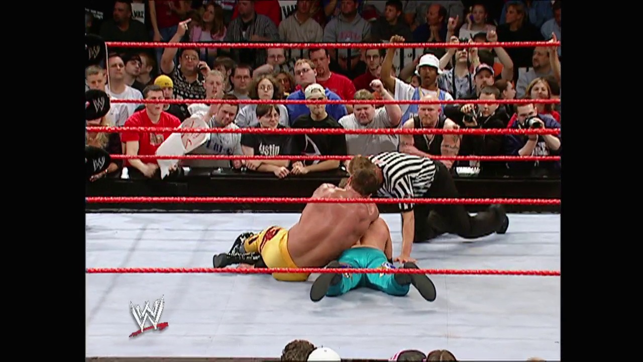 A frame from WWE Raw (November 15, 2004) at 54:37. A WWE wrestler lies pinned on the mat while a referee in a striped shirt counts, with the crowd visible behind the red ropes and the WWE logo displayed in the corner.