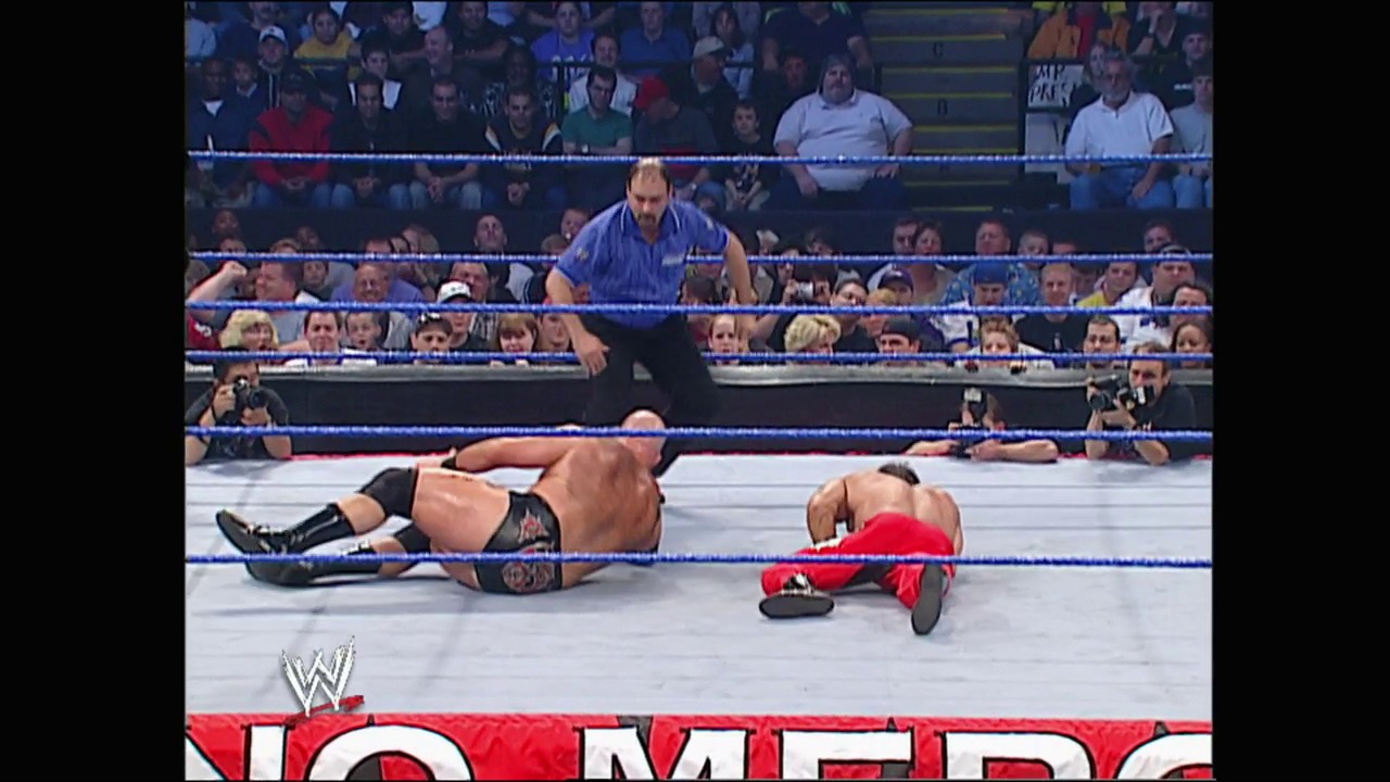 A frame from WWE No Mercy (2003) at 24:00. Two wrestlers lie on the mat during a WWE match, with one referee standing over them. The crowd fills the arena seats while the WWE logo and "No Mercy" branding are visible at the bottom of the screen.