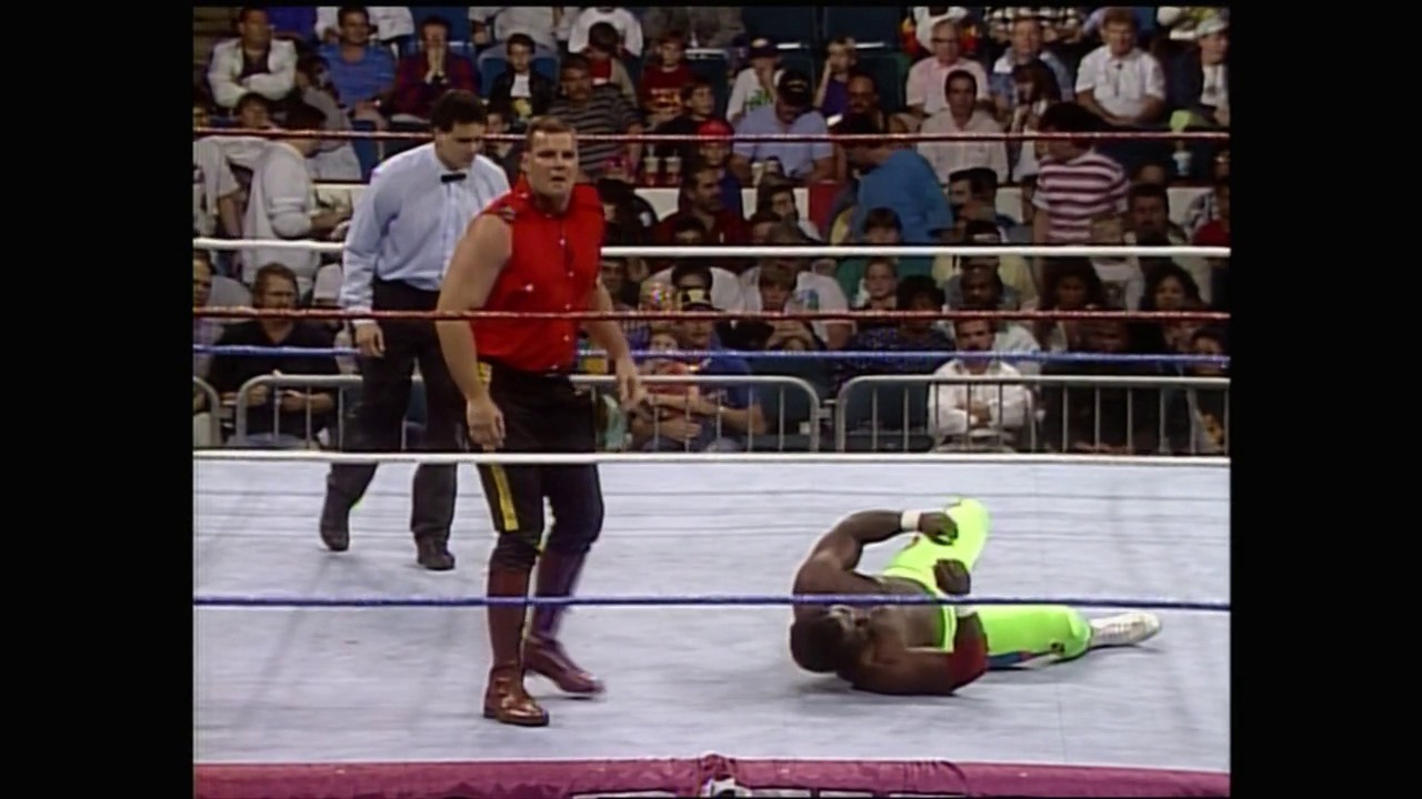 A frame from WWE Royal Rumble 1991 (1991) at 1:20:33. A wrestler in neon green lies on the mat while an opponent in red stands nearby during a WWE Royal Rumble 1991 match, with a full crowd visible in the background and officials at ringside.