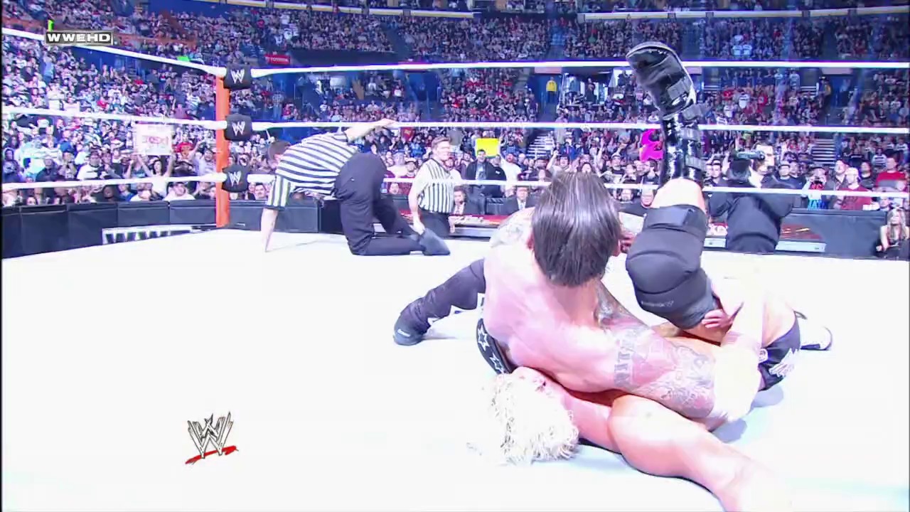 A frame from Royal Rumble (2012) at 1:32:38. A WWE wrestler in pink attire is pinned on the mat during a Royal Rumble match, with a referee in striped shirt overseeing the action. The packed arena crowd fills the background behind the white wrestling ring.