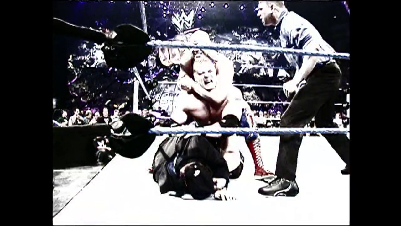 A frame from WrestleMania XIX (2003) at 3:13:24. # WrestleMania XIX Frame

A wrestler in white lies on the mat while another competitor stands nearby with a steel chair, as a referee observes the action. The crowd is visible in the background of the professional wrestling ring.