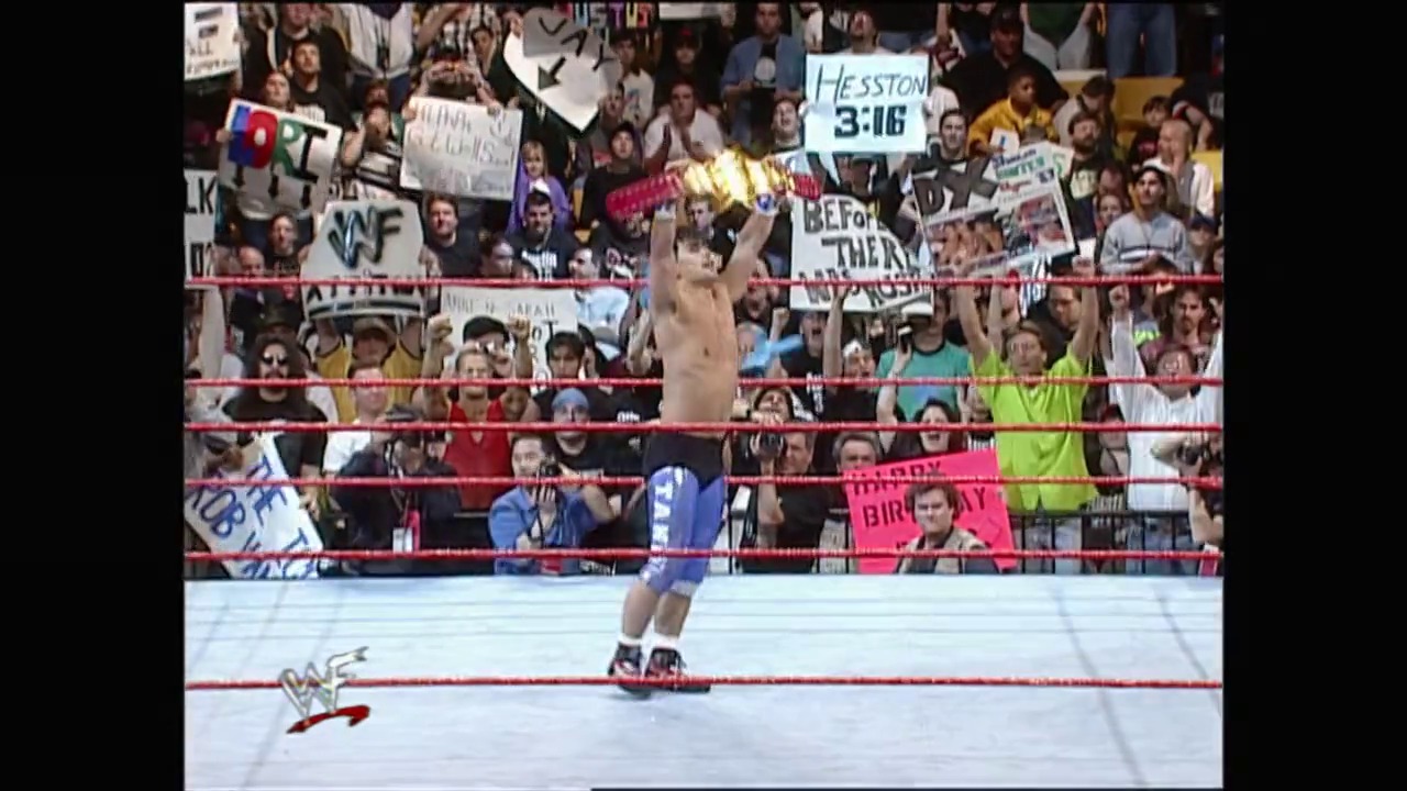 A frame from WrestleMania XIV (1998) at 24:04. A shirtless wrestler in blue shorts celebrates in the WWF ring, holding a championship belt aloft while the crowd cheers from behind the red ropes. Various fan signs are visible in the packed stadium during this iconic WrestleMania XIV moment.