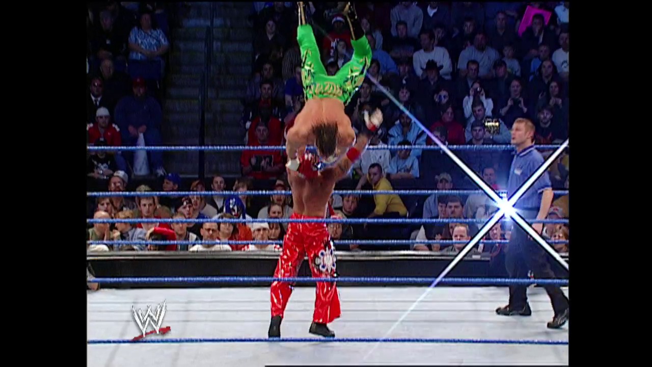 A frame from WWE Raw (November 14, 2002) at 34:42. A WWE wrestler performs an aerial maneuver from the top rope, suspended upside-down in a bright green and yellow outfit, while an opponent in red attire stands below in the ring. The blue-roped WWE ring is surrounded by an enthusiastic crowd, with the WWE logo visible in the corner.