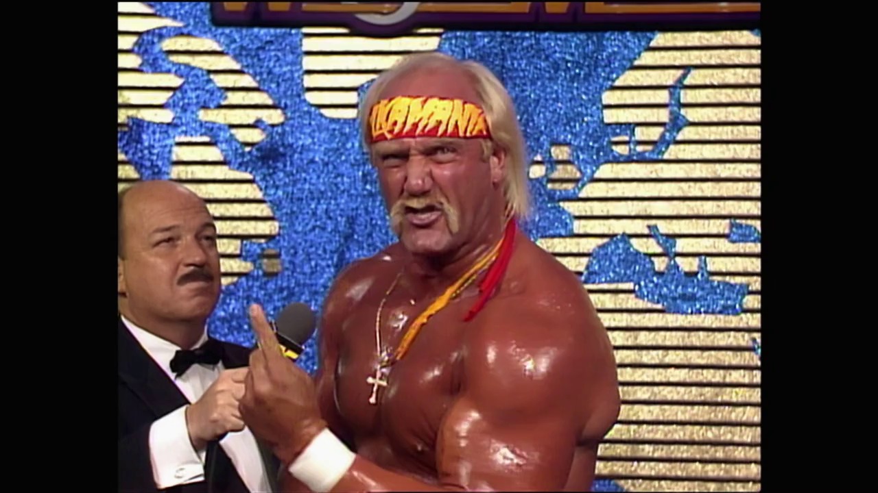 A frame from WrestleMania IV (1988) at 1:11:03. A professional wrestler in red and gold attire with a yellow headband stands at a microphone during WrestleMania IV, while an interviewer in formal wear holds the microphone. The blue and gold set design with horizontal striping is visible in the background.